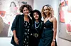 Nafkot Gebeyehu, Mahlet Afework, and Zion Yaynu standing together at Studio 11 during the opening of Frozen Moments, Afework's solo exhibition. The trio is smiling in front of colorful paintings that capture moments of Ethiopian life.