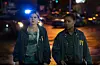 Two women in FBI jackets walk through a dimly lit street at night, looking serious. One carries a folder and the other has a gun holstered at her waist. Police vehicles with flashing blue lights are visible behind them.