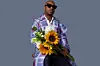 Da Capo holds a bouquet of sunflowers in a promo image for his latest album. He is wearing an elegant purple coat by Thebe Magugu, navy blue pants that split open at the feet, and a white shirt. He is also wearing dark shades.