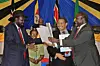 South Sudanese President Salva Kiir, wearing a dark suit, red tie, and wide-brimmed black hat, shakes hands and exchanges documents with opposition leader Riek Machar, also in a dark suit and red tie, as they smile during a peace agreement ceremony. Former Tanzanian President Jakaya Kikwete stands between them, grinning and holding papers, while other officials and colorful East African flags form the backdrop.