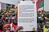 A crowd of demonstrators, mostly men, gather closely together waving Burkina Faso flags and wearing patriotic clothing. One man in the foreground holds a large placard with a French message criticizing Western influence and calling for African unity and sovereignty. The atmosphere is lively and energetic, with vuvuzelas, signs, and national pride on full display.