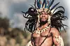 A still from 'Shaka iLembe' showing actor Thembinkosi Mthembu dressed in traditional warrior attire, featuring a feathered headdress and necklace with large bone pieces.