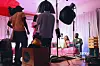 Behind-the-scenes of a video shoot, with crew members adjusting lighting and equipment while two people relax on a pink couch in the background.