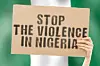 The phrase “Stop the violence in Nigeria” on a banner in a man’s hands, with a blurred Nigerian flag in the background.