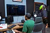 Two people in a music studio, one seated at a desk using a keyboard and production software on multiple screens, while the other, wearing a bright green face mask, stands next to them, observing the process.