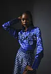 A model poses for a photo in a dimly-lit room wearing a blue shirt tucked into a blue skirt, both made from Akwete fabric.