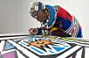 Iconic South African artist Esther Mahlangu, adorned in traditional Ndebele attire and intricate beadwork, leans over a large canvas as she carefully paints a vivid geometric pattern in blue, orange, black, and white. Her silver hair is styled in a crown-like shape and decorated with beads, and she wears colorful layers of fabric and jewelry, embodying the rich heritage of her culture.