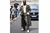 \u200bAdekunle Gold wears silver earrings, white gold diamond choker necklace, white buttoned up long sleeve shirt, dark brown trench coat, dark green tote bag, dark olive green baggy pants, black sunglasses in hand, light yellow gemstone ring, white black Bluemarble loafers shoes