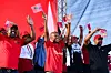 EFF president Julius Malema, in a red t-shirt, waves the flag of his party while smiling, surrounded by members of his party, also waving flags.