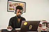 A Black man with a short afro and goatee stits at his desk looking at an open laptop.