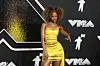 A young woman poses for the cameras on the VMA red carpet. She is wearing a tight mini dress in neon yellow, with slits at the front that reveal neon green underwear. The woman is also wearing a bright blue choker-style necklace with a honey-blonde Afro with loose curls.
