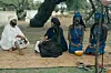 A group of people sits on a mat under a tree \u2014 a man speaking to a woman, and two women looking downward beside them.