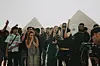 Egyptian rappers gathered in front of the Great Pyramids of Giza, one masked man is holding a microphone to a young woman rapping in the middle of the group.