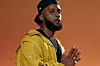 Maleek Berry stands in front of a beige background, looking at the camera sideways.