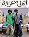 Ahmed Atef and Mohamed Tarek standing in front of the plastic chairs of a coffee shop, wearing wide jeans and flowy tops in green and blue, looking straight at the camera.