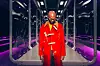 South African rapper, the late Riky Rick, poses for the camera. He is bald, wearing a shiny red coat and large sunglasses standing on a futuristic walkway lit with purple lights, looking down.