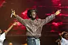 A photo of Burn Boy on stage, smiling and wearing a brown jacket and a cowboy hat.