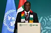 Emmerson Mnangagwa, President of Zimbabwe, wearing a colorful scarf in Zimbabwe’s national colors, is speaking at a podium with United Nations and COP28 UAE logos, flanked by UN and national flags