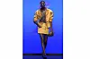 A tall, dark-skinned woman in a yellow outfit is seen walking down a runway.