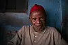 An elderly man wearing a bright red cap sits staring at the camera.