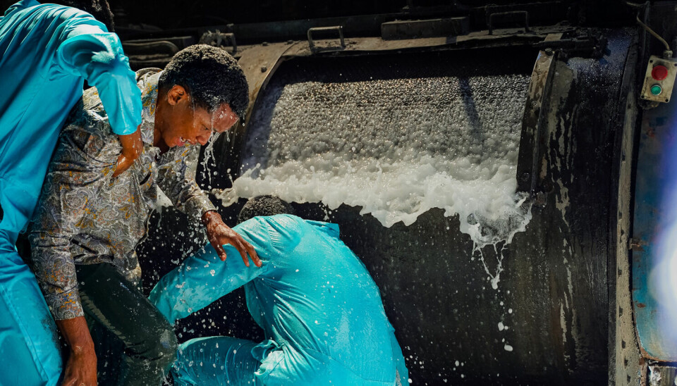 Zamo Mkhwanazi’s ‘Laundry’ is a potent period drama that lays bare the challenges and complexities of existing under a brutally racist regime. A still from Zamo Mkhwanazi's 'Laundry' showing two men in blue uniforms holding a soaked man in a patterned shirt near a large industrial washing machine overflowing with water and foam.