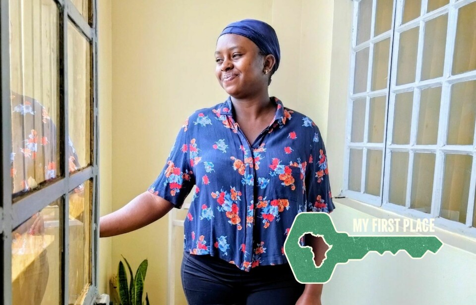 Kenyan chef and food stylist Geena Gichui at home in Kiambu, where she is adjusting to life outside Nairobi and embracing the independence of her first apartment. A woman wearing a floral shirt and black shorts stands smiling inside her apartment near a doorway, with a sofa, curtains, and a potted plant visible in the background.