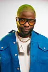 Skales wears a blue jacket over a white shirt, topped with colored sunshades; his hair is dyed green.