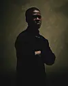 A profile image of Nasty C. He wears a black shirt, is folding his hands, his face is turned towards the camera, and deep shadows cover one side of his face.