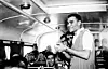 Black and white photograph of a man standing in the aisle of a bus full of young men who are singing or yelling. He is smiling.
