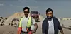 Two men, one wearing the outfit of a construction worker and the other a simple button-down shirt, are walking with grim faces, as if they’re on a mission, on a construction site.