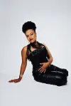 Ethiopian singer and producer Meklit poses for a photo. She’s wearing a black romper jumpsuit, and she’s seated in an all-white background.
