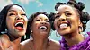 Three women huddled together with big smiles, the sky in the background