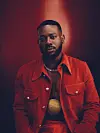 Adekunle Gold looks straight at the camera. He wears a red outfit, no shirt underneath, a silver chain, and a neck piece with a large, round gold pendant.