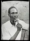A medium close-up black and white self-portrait of the artist looking at the camera with a flower in his hand, brought up to his closed mouth.