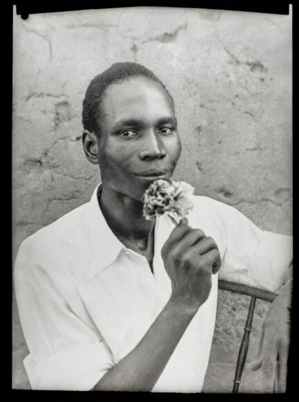 Seydou Keita. Untitled, 1956, printed 2018. A medium close-up black and white self-portrait of the artist looking at the camera with a flower in his hand, brought up to his closed mouth.