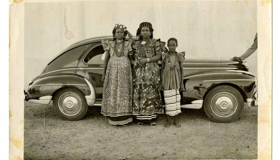 Seydou Keita. Untitled, 1954. A sepia-toned photograph of three women of different ages standing in front of an old car, wearing dresses. The photographer’s image can be seen in the reflection of the car bonnet.