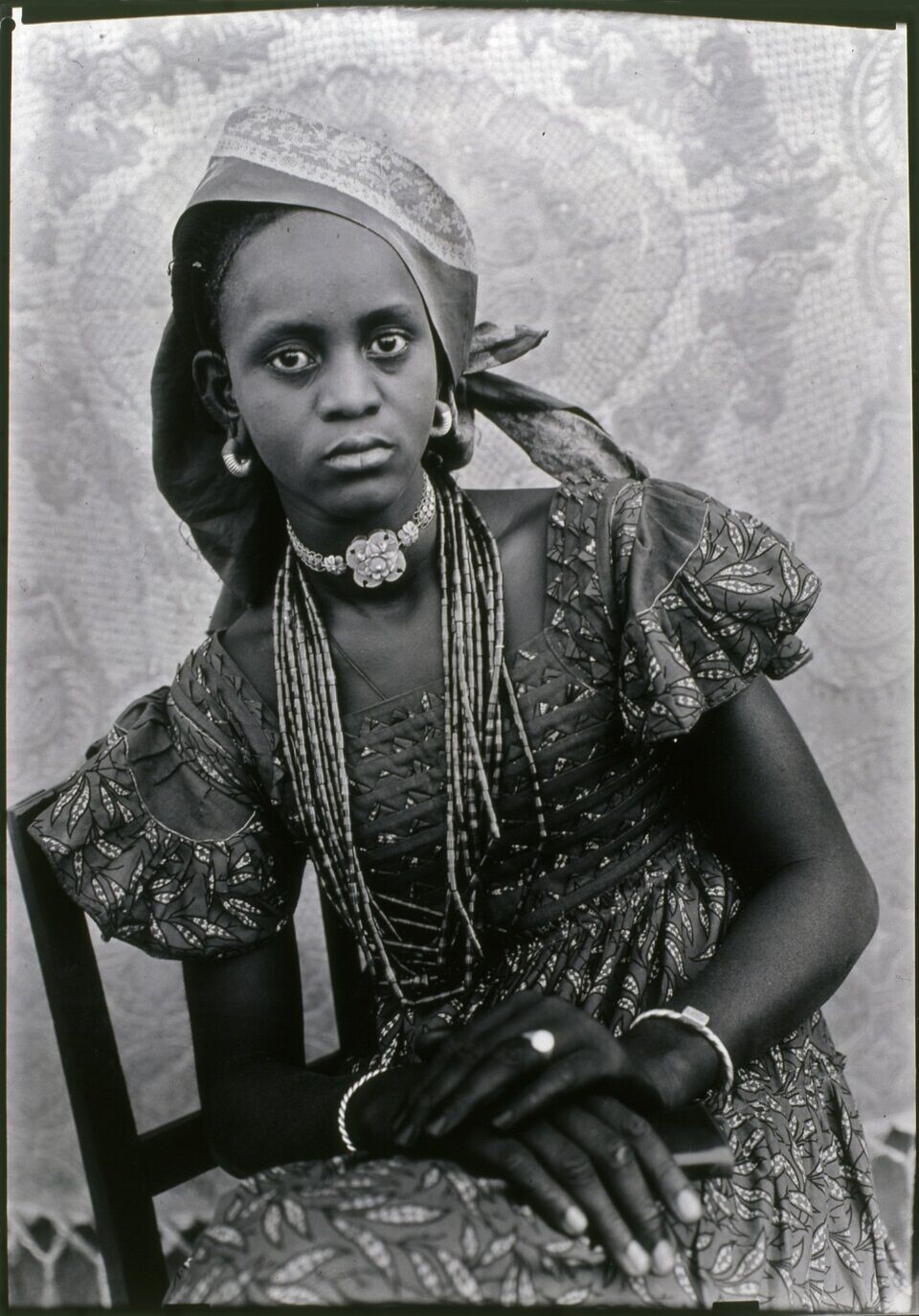 Seydou Keita. Untitled, 1956, printed 2018. A medium close-up black and white portrait of a woman looking at the camera, wearing jewelry, with her hands crossed over her lap.