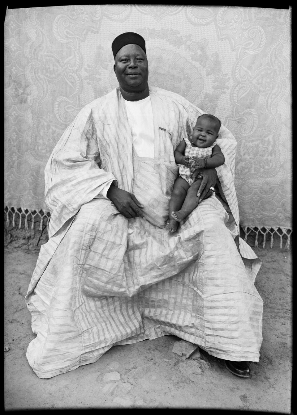 Seydou Keita. Untitled, 1949-51, printed 1998. A man in a boubou is holding a baby who is wearing an outfit in a similar textile, and both are smiling widely.