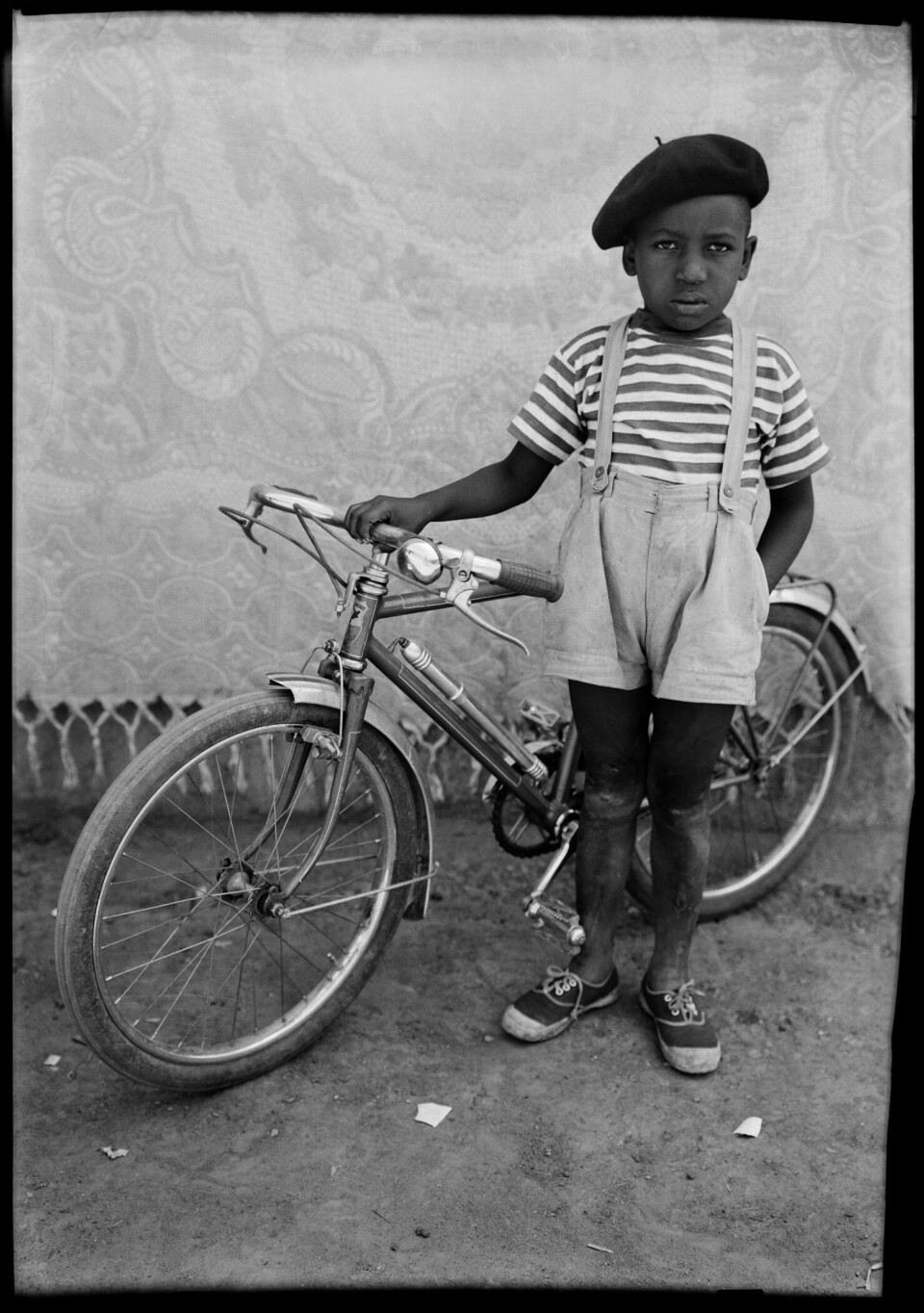 Seydou Keita. Untitled, ca. 1949-51, printed 1995. A little boy is standing in front of a bicycle with a solemn look on his face, and he’s wearing a beret and short overalls.