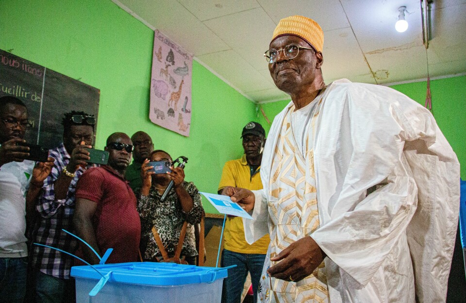 Cameroon opposition figure Issa Tchiroma Bakary casts his ballot on Sunday, October 12, 2025. He declared victory in the presidential election today. Issa Tchiroma Bakary smiles as he gets ready to cast his ballot. People around him stand with their phones and cameras, taking pictures.