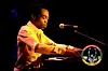 Fela Kuti is on stage in front of an instrument and a microphone, wearing a yellow shirt