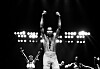 A black-and-white picture of musician and composer Fela Anikulapo-Kuti posing while on stage in Detroit, Michigan, in 1986. Kuti stands shirtless with both fists raised, his saxophone strap hangs around his neck and behind him are some of the female dancers and backup singers that often performed alongside him.