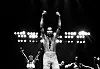 A black-and-white picture of musician and composer Fela Anikulapo-Kuti posing while on stage in Detroit, Michigan, in 1986. Kuti stands shirtless with both fists raised, his saxophone strap hangs around his neck and behind him are some of the female dancers and backup singers that often performed alongside him.