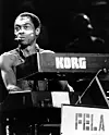 A black-and-white picture of Fela Kuti on stage at London’s Brixton Academy in 1989. Kuti is seated in front of a piano with a cigarette stick in his mouth.