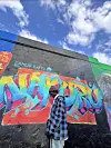 A man in a plaid blue jacket and a beanie stands beside a colorful graffiti mural that spells “Nakuru” in bold letters on a black wall