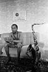A black-and-white picture of Fela Anikulapo-Kuti. He’s sitting on a three-seater couch wearing only briefs, a saxophone strap hangs around his neck, and a saxophone rests on the couch beside him.