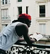 Stonebwoy stands on the balcony looking out to the street in a promo shot. He is wearing a red beret, dark shades, and a white vest.