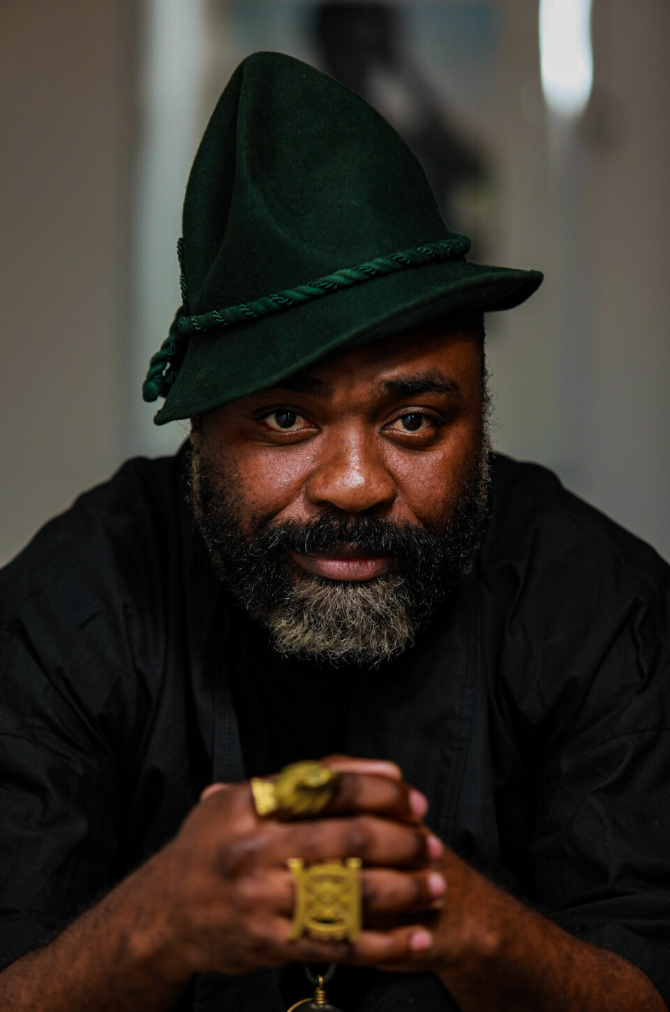 Makhathini did not set out to become a jazz musician, and now he is a master at his craft, celebrated globally. Nduduzo Makhathini wears a green hat and a black shirt. His hands are clasped in front of him, fingers in large gold rings, and he stares ahead at the camera.