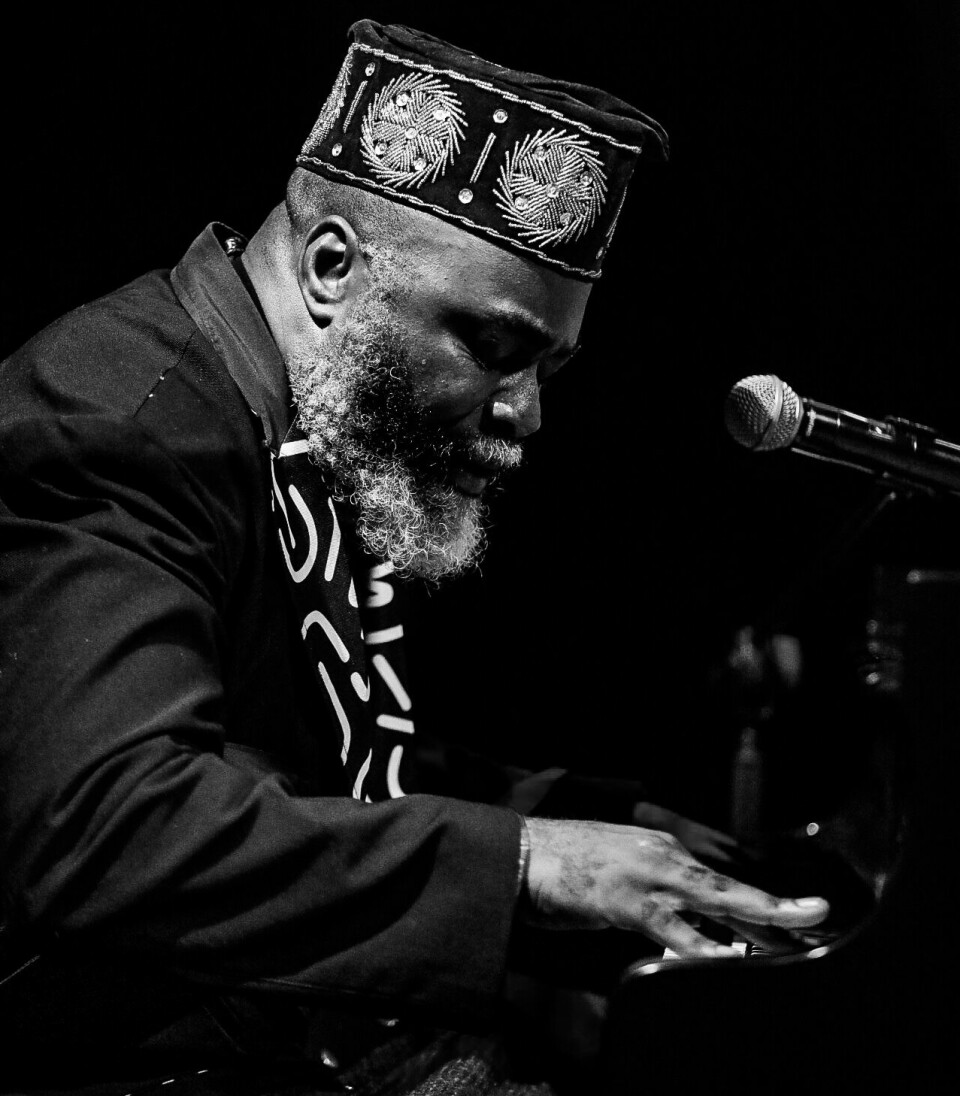 “Sound tells people how to experience it. It defaults to something greater than race or place.” A black and white image of Nduduzo Makhathini playing a piano, a microphone in front of him. He wears a black kofia with geometric beadwork that reflects cosmological and ancestral meanings.