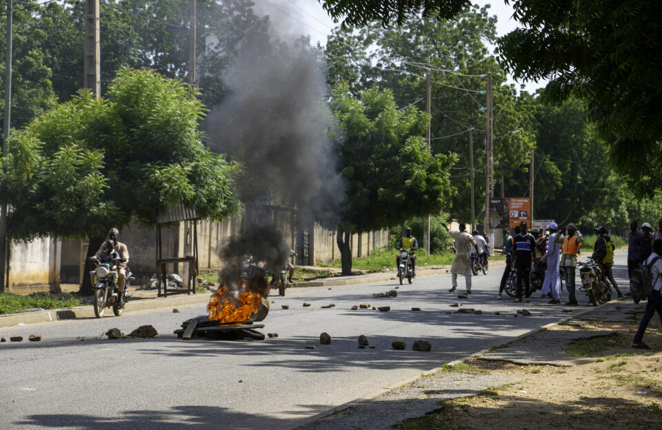 Burning barricades are seen in Garoua during a demonstration by supporters of the political opposition on October 21, 2025, ahead of the release of the results of the presidential vote. A burning tire sits in the middle of a street, while a group of protesters is seen in the background, and a motorbike is passing through.